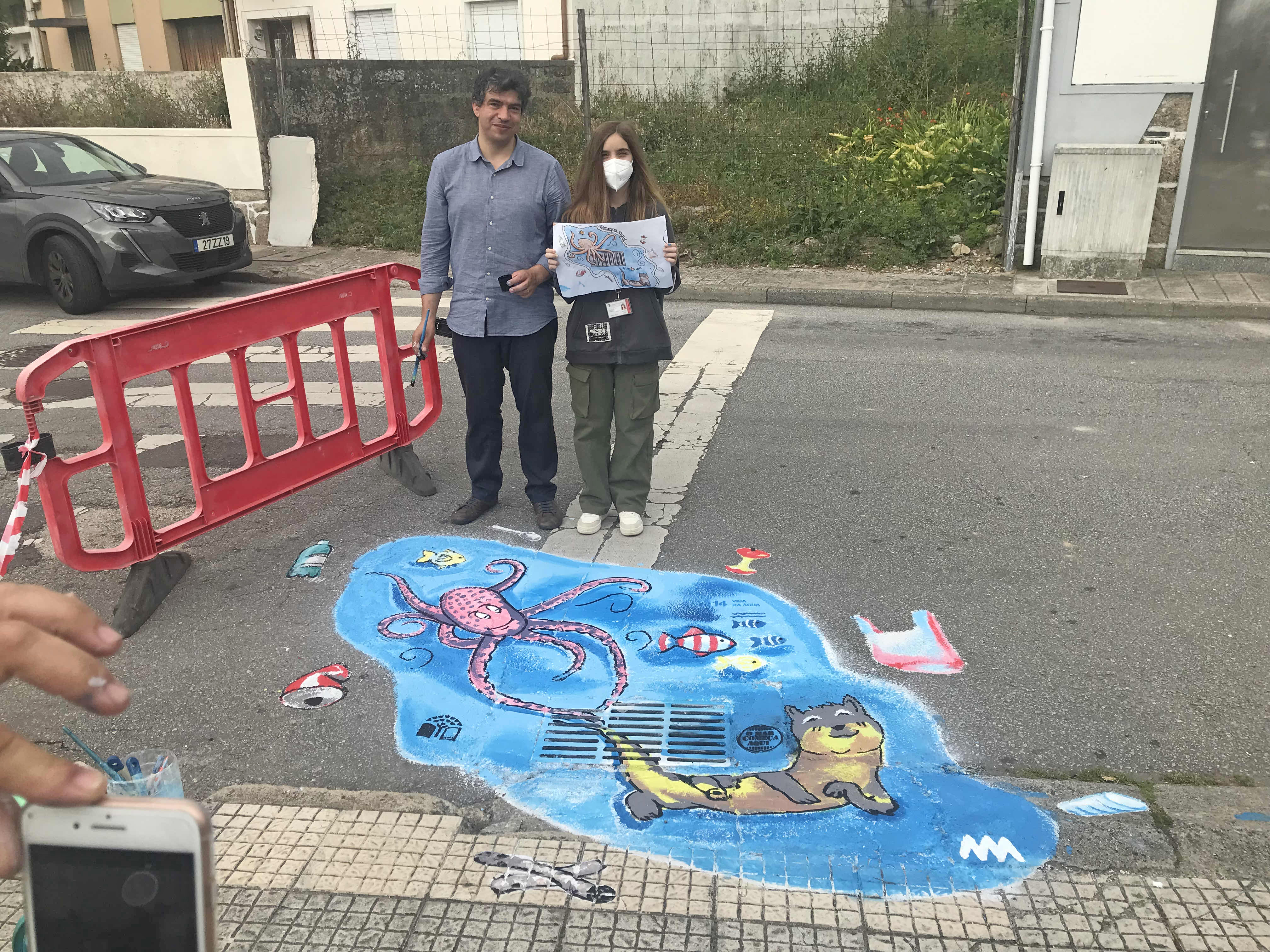 Gabriela Cortes, a segurar a imagem da sua ilustração junto ao professor de Educação Visual e do trabalho executado junto da grelha de escoamento de águas pluviais.
